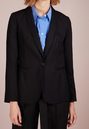 Blazer noir avec fermeture à un bouton, revers crantés et deux poches avant. Porté sur une chemise à col bleu clair. Texture de tissu lisse.