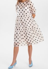Lätt, genomskinlig klänning med prickar i svart på en krämfärgad bakgrund. Har puffärmar och en lager-på-lager, svävande design. Blå klackar.