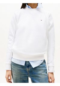 Sudadera blanca con cuello redondo, puños acanalados, con un pequeño logo. Lleva puesta sobre una camisa de botones azul clara y jeans azules.