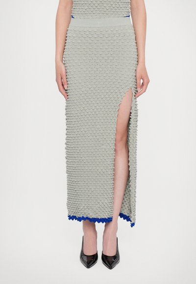 Robe maxi tricotée en gris clair avec des motifs texturés en relief, une fente jusqu'à la cuisse et un ourlet en dentelle bleue. Associée à des talons noirs.