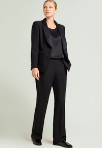 Schwarzer Hosenanzug mit einem maßgeschneiderten Blazer mit Satinkragen, gerade geschnittene Hosen und einem seidigen schwarzen Top, alle aus einem glatten Stoff.