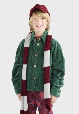 Chemise en velours côtelé verte avec des boutons en bois, t-shirt rouge en dessous, casquette bordeaux, et écharpe grise/rouge sur un pantalon de pyjama à motifs.