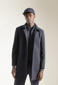 Abrigo de mezcla de lana en color navy con solapa, botones frontales y bolsillos laterales. Lleva puesto un chaleco acolchado oscuro y se complementa con una gorra a juego.