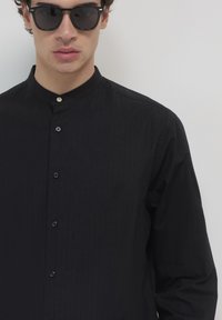 Schwarzes Hemd mit Knopfleiste und gestreiftem Muster, Mandarin-Kragen und kontrastierenden Knöpfen. Getragen mit dunklen Sonnenbrillen. Glatte Stoffstruktur.