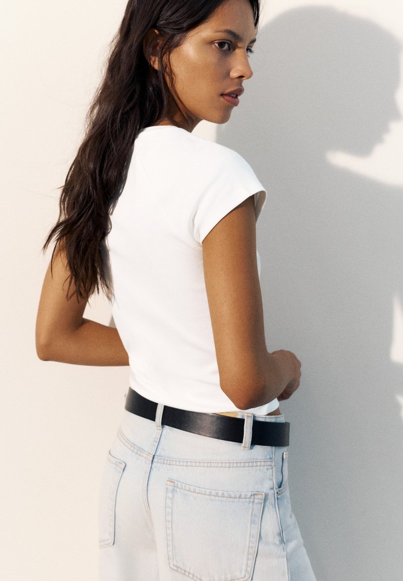 Femme aux longs cheveux foncés portant un haut blanc à manches courtes et un jean bleu clair avec une ceinture noire, regardant par-dessus son épaule.