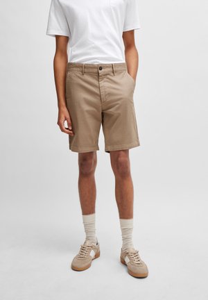 Pantaloncini beige realizzati in tessuto leggero, con chiusura frontale a bottone, tasche laterali e una vestibilità casual. Abbinati a sneakers chiare e calzini.