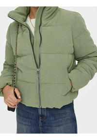 Chaqueta verde de pana con diseño abullonado, cuello alto, cierre de cremallera y bolsillos laterales. Se lleva sobre una camiseta clara y combinada con vaqueros.