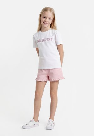 Ung pige står, iført en hvid Missoni t-shirt, lyserøde flæse-shorts og hvide sneakers, smilende til kameraet.