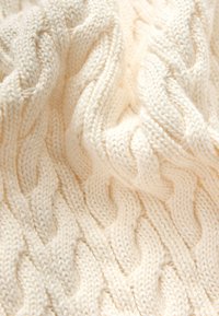 Kremų spalvos megztinis su pintinio raštu, turintis banguojančių bangų ir iškilusių dygsnių tekstūrą, pagamintas iš minkštos siūlų medžiagos.