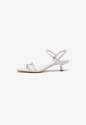 White strappy sandal featuring a low block heel, flat sole, and adjustable ankle strap. Smooth material with a simple design.
