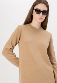 Maglione beige a girocollo con maniche lunghe, morbido texture a maglia e design minimalista, indossato con occhiali da sole neri angolari.