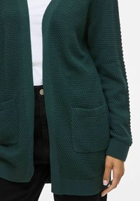 Zeleni pleteni kardigan z zanimivim vzorcem, odprt spredaj, z dvema sprednjima žepoma in rebrastimi manšetami. Enostaven in vsestranski dizajn.