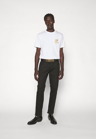 Camiseta blanca de manga corta con un gráfico de un oso, combinada con pantalones negros y un cinturón dorado con el logo "MOSCHINO". Zapatos de vestir negros completan el conjunto.