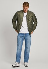 Chaqueta bomber verde oliva con textura suave, bolsillos con cremallera, combinada con una camiseta blanca y jeans azules. Zapatillas deportivas completan el look.