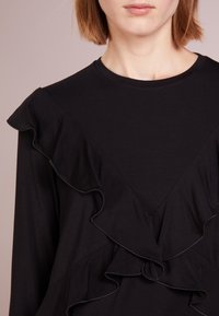 Svart långärmad topp med volangdetaljer längs halsringningen och bodice, tillverkad av mjukt tyg med en slät textur.