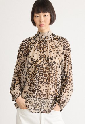 Vrouw in een blouse met lange mouwen en hoge hals in luipaardprint, met elastische manchetten, met handen in de zakken, tegen een plain witte achtergrond.