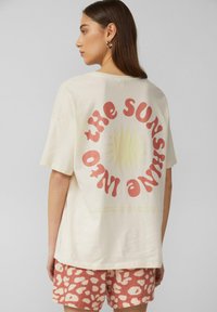 Crème kleurige katoenen t-shirt met een grote rode en gele zonafbeelding en tekst op de achterkant, gecombineerd met roze gemerkte shorts. Korte mouwen en een relaxte pasvorm.