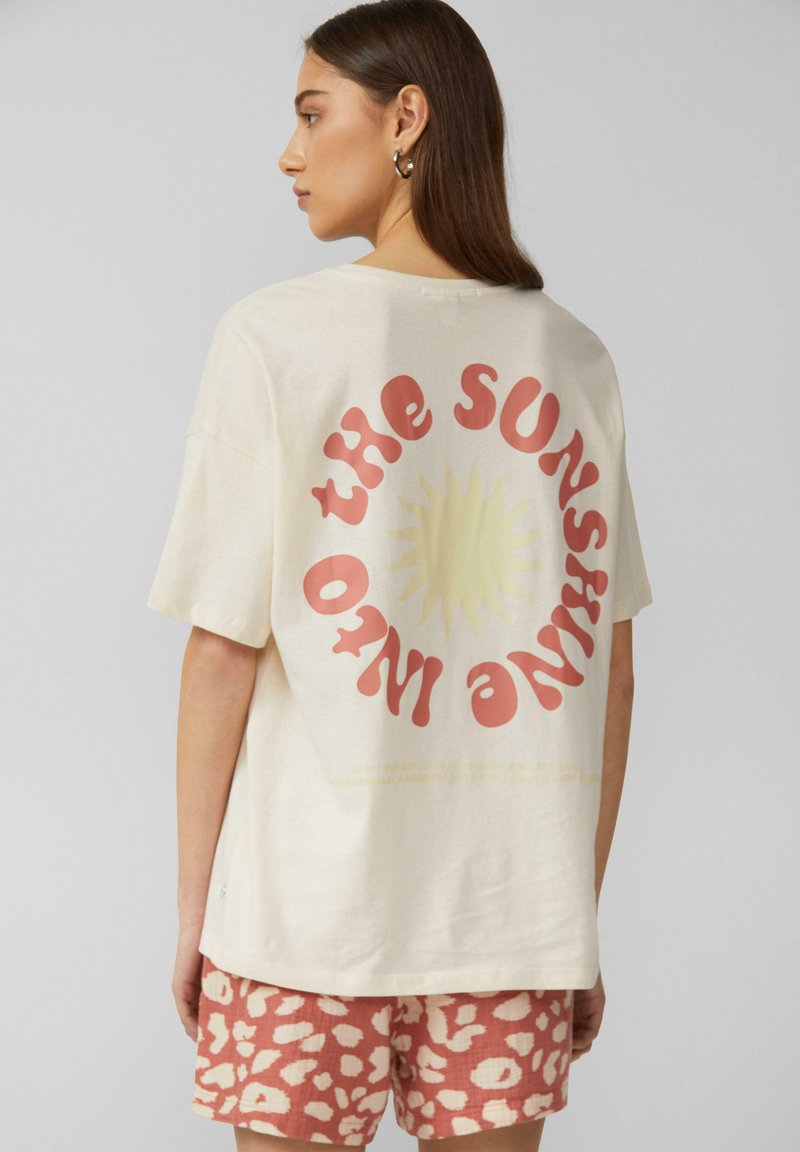 Crème kleurige katoenen t-shirt met een grote rode en gele zonafbeelding en tekst op de achterkant, gecombineerd met roze gemerkte shorts. Korte mouwen en een relaxte pasvorm.