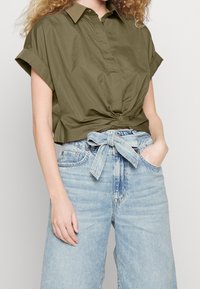 Camicia corta verde oliva con colletto e dettaglio antracite all'anteriore, abbinata a jeans in denim leggeri blu chiaro a vita alta con cintura da annodare.