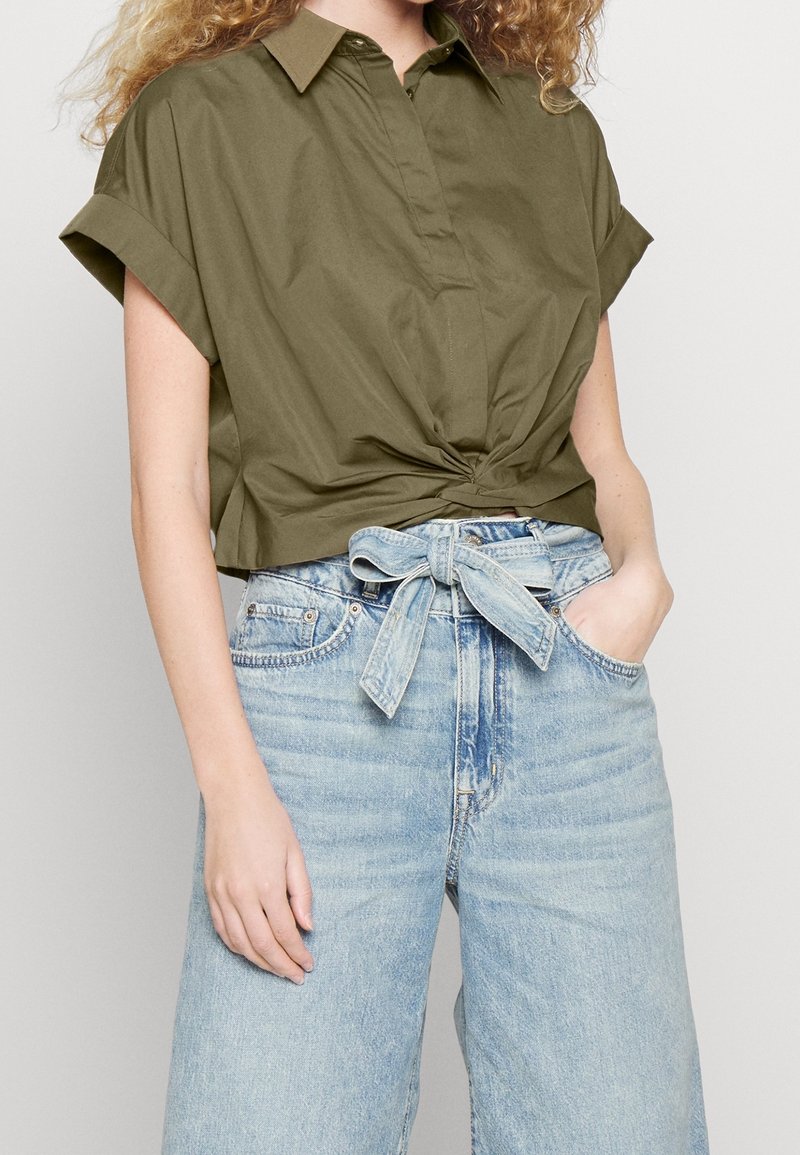 Camicia corta verde oliva con colletto e dettaglio antracite all'anteriore, abbinata a jeans in denim leggeri blu chiaro a vita alta con cintura da annodare.