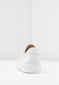 Baskets blanches en cuir au toucher lisse, avec un talon arrondi et une semelle en caoutchouc plate. Elles présentent un logo discret et un design minimaliste.