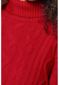 Roter Strickpullover mit hohem Kragen, Zopfmusterstruktur und aufwendigen Mustern, die verschiedene Maschen und Stärken hervorheben.