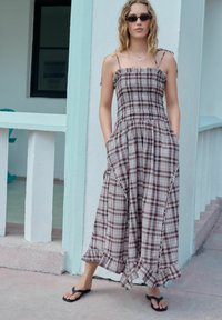 Maxime dress with a plaid pattern in reds, browns, and grays. Features a smocked bodice, spaghetti straps, and a ruffled hem. Worn with flip-flops.