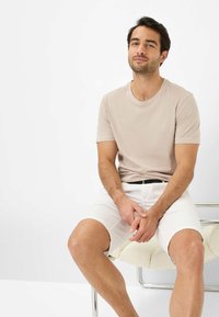 Beige short-sleeve t-shirt with a round neck, paired with white shorts. Sitting on a chair with a textured seat. Neutral background.