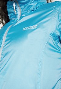 Veste imperméable bleu clair avec fermeture zippée à l'avant, accents réfléchissants et logo sur la poitrine. Tissu lisse et léger.