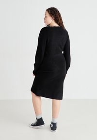 Vestido de punto negro con mangas largas, cintura ajustada y dobladillo a la altura de la rodilla. Combinado con zapatillas altas negras y calcetos grises con estampado.
