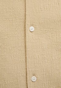 Camisa texturizada beige con un patrón arrugado y dos botones blancos. La tela parece ligera y transpirable.