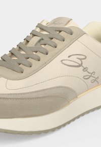 Basket beige et gris clair avec semelle texturée, lacets gris, détails en daim et logo "Boss" en cursive sur le côté et la languette.
