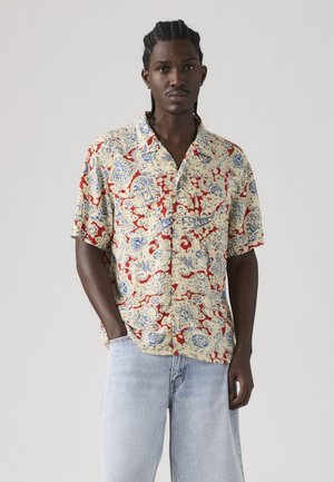 Homme aux cheveux tressés portant une chemise à manches courtes boutonnée avec des motifs abstraits rouges, bleus et beiges et un jean bleu clair, debout sur un fond gris.