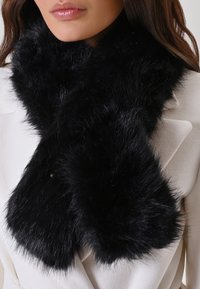 Écharpe en fausse fourrure noire avec une finition douce et texturée, présentant des bords arrondis et un accent doré caché. Portée par-dessus un manteau de couleur claire.
