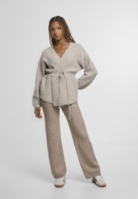 Jasnobeżowy kardigan typu wrap z dekoltem w serek, wiązany w talii, z szerokimi rękawami, zestawiony z pasującymi spodniami o szerokich nogawkach, wszystko w miękkiej dzianinowej fakturze.