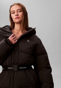 Young woman wearing a black Calvin Klein puffer jacket with a hood, adjusting the collar, against a plain gray background.