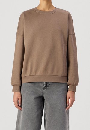 Persoon draagt een losse bruine crewneck-trui met lange mouwen en omgeslagen polsen, gecombineerd met grijze jeans met hoge taille.