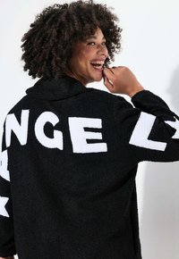 Veste noire en fourrure douce avec un grand texte blanc à l'arrière. Comprend un col et un tissu doux et texturé. Le motif inclut des lettres graphiques et audacieuses.