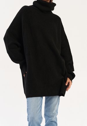 Femme portant un pull noir à col roulé oversize avec épaules tombantes et jean denim bleu clair sur fond uni.