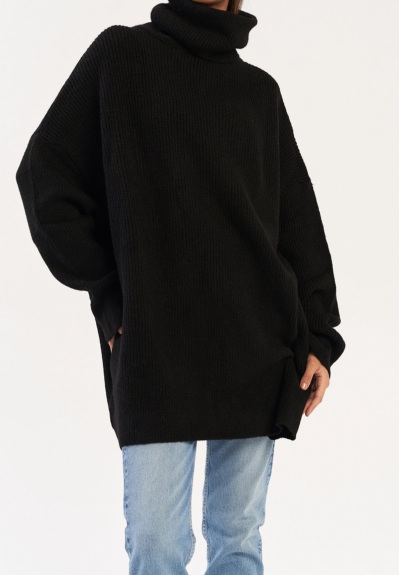 Femme portant un pull noir à col roulé oversize avec épaules tombantes et jean denim bleu clair sur fond uni.