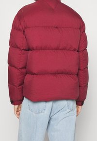 Bordeaux pufferjack met verticale, gewatteerde secties, een hoge kraag en elastische manchetten. Draagt over lichtblauwe jeans met zichtbare achterzakken.