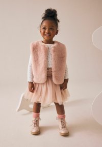 Un gilet en fausse fourrure rose sur une chemise à manches longues à motifs floraux clairs, associé à une jupe en tulle et des bottes roses avec des chaussettes assorties.