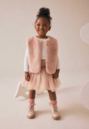 Jeune fille souriante portant un gilet en fausse fourrure rose, une chemise blanche à manches longues, une jupe en tulle rose, des chaussettes roses et des bottines à lacets rose clair.