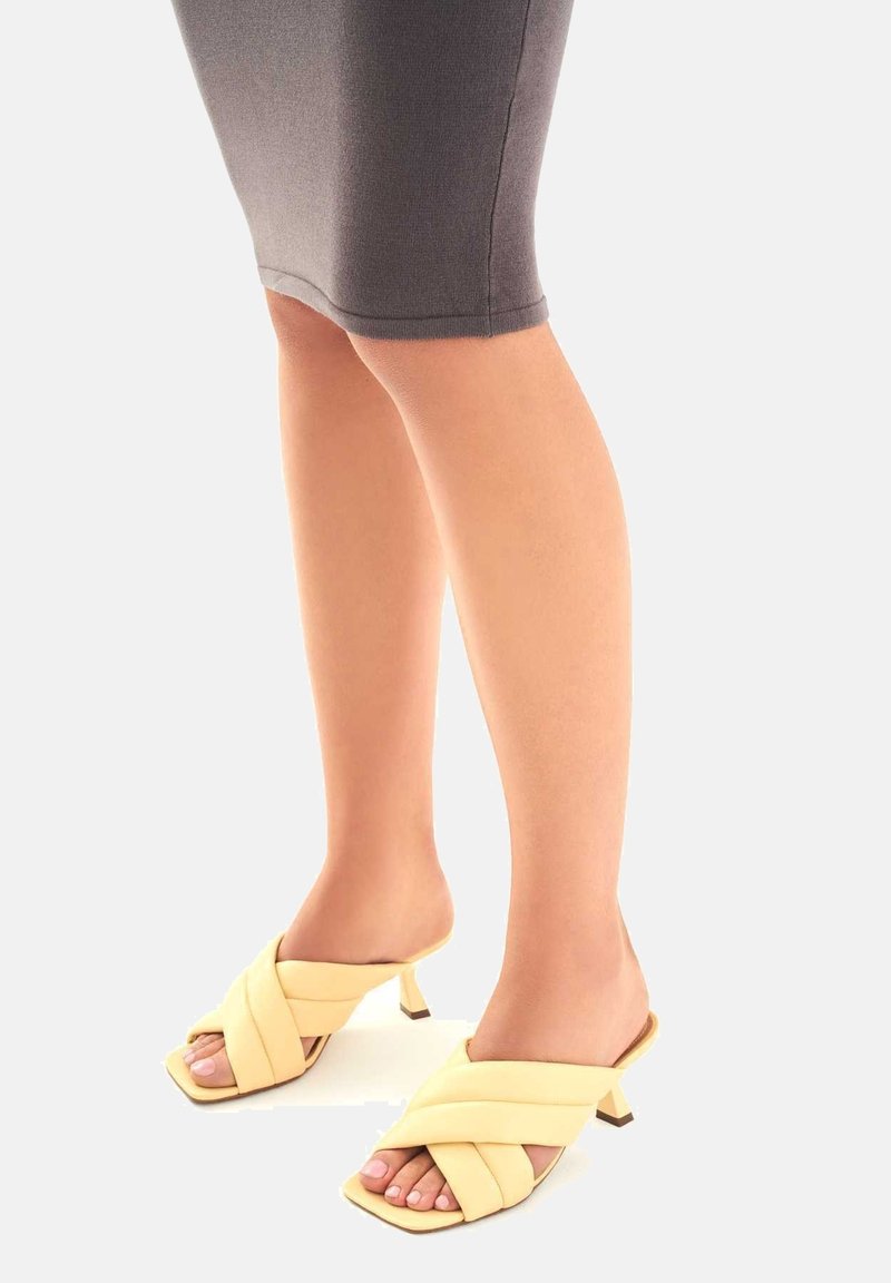 Mules à lanières croisées jaunes avec un petit talon et un bout ouvert. Texture lisse avec un design moderne, sur fond gris clair.