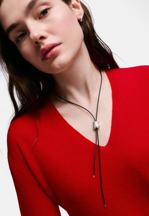 Joven mujer con cabello oscuro que lleva un suéter rojo de cuello en V y un collar de cuerda negra con un colgante de osito de plata.