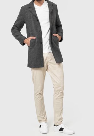 Homme portant un manteau gris boutonné, un t-shirt blanc, un pantalon beige et des baskets blanches sur un fond uni gris clair, mains dans les poches du manteau.