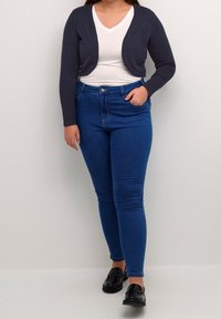 Schwarze Strickjacke, weißes V-Ausschnitt-T-Shirt, blaue Skinny-Jeans und schwarze Slipper. Weiches Strickmaterial, figurbetonter Schnitt, mittelhoher Taillenbund und glatte Textur.