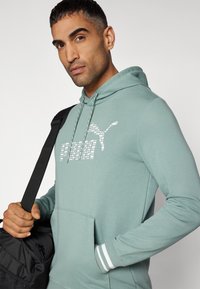 Felpa verde chiaro con cappuccio, tasca frontale, polsini a strisce bianche e un design del logo Puma riflettente. Il tessuto appare morbido e liscio.