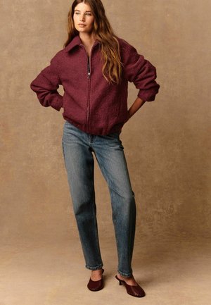 Jeune femme se tenant les mains sur les hanches, portant une veste zippée bordeaux, un jean bleu et des mules à talons bordeaux, sur un fond neutre.