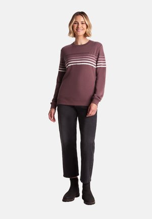 Smiling woman with short blonde hair wears a purple striped sweatshirt, black cropped jeans, and black ankle boots, standing on white background.